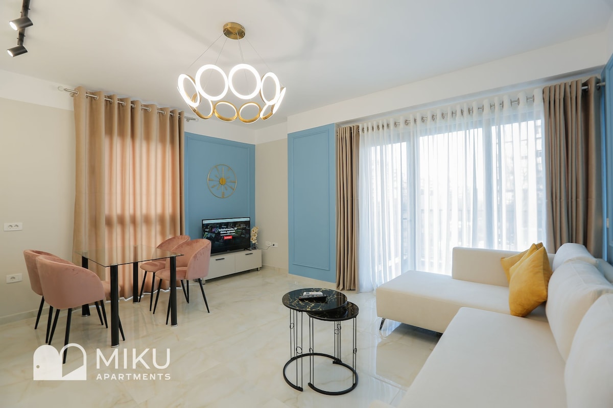 A modern living area displays a white sectional sofa paired with a decorative pillow. A circular chandelier hangs above a glass coffee table set with two black dining chairs. Natural light filters through sheer curtains, highlighting a simple entertainment unit and decorative wall clock.