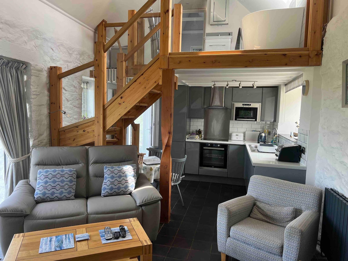 An open-plan living area is highlighted by wooden beams and a staircase leading to an upper level. Comfortable seating is arranged around a coffee table, and a modern kitchen is visible in the background, featuring sleek appliances and ample counter space.