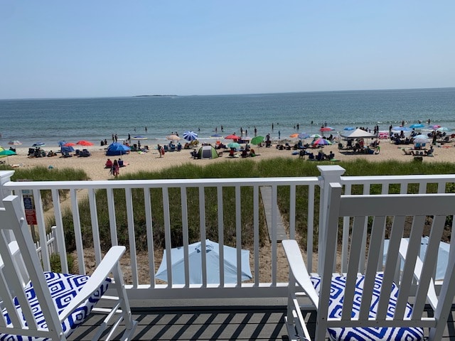 The private deck offers a serene view of the beach and ocean, where umbrellas and beachgoers can be seen enjoying the sand. Two inviting chairs with blue and white patterns are positioned for relaxation while taking in the coastal scenery.