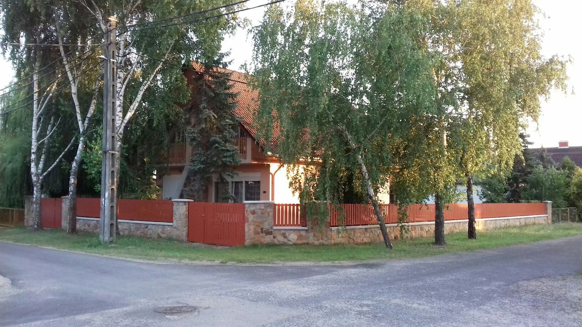 The two-story house is framed by lush greenery, featuring a stone exterior and a red wooden fence. The surrounding area is tranquil, showcasing a grassy lawn and trees that provide shade. The building's roof is gabled, enhancing its charming appearance.