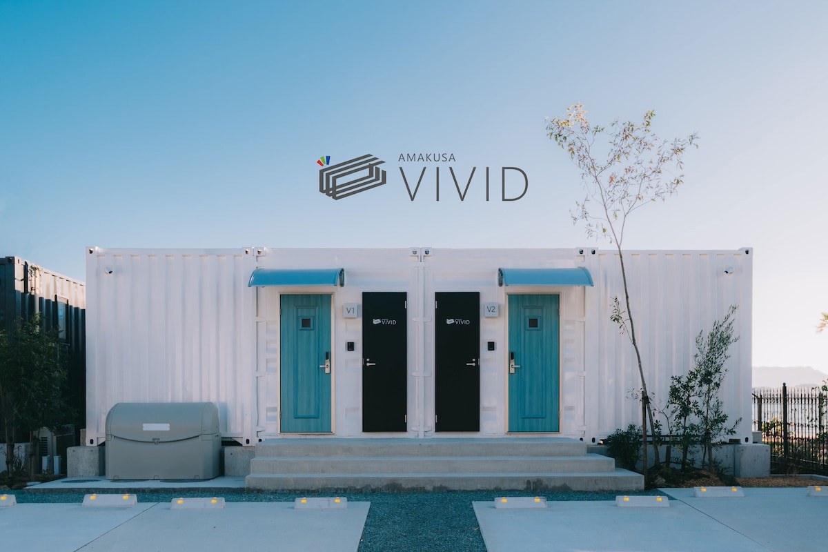 A modern container house is positioned along a coastal location, featuring two distinct entrances with contrasting doors in black and turquoise. A concrete pathway leads to the doors, while subtle landscaping is present in the surrounding area.