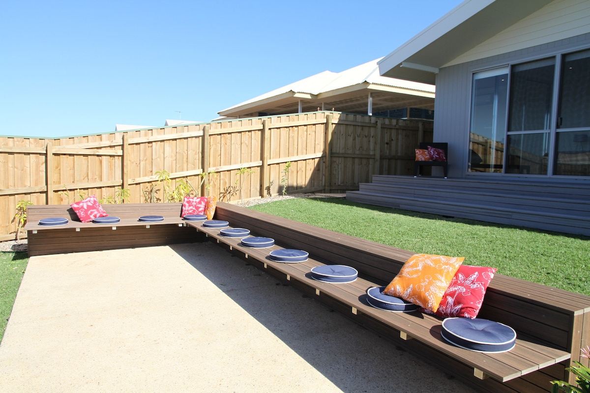 An outdoor seating area is shown with built-in benches, featuring colorful cushions. Round plates are arranged on the surface, inviting casual dining. The space is bordered by a well-maintained lawn and fencing, under a clear blue sky.