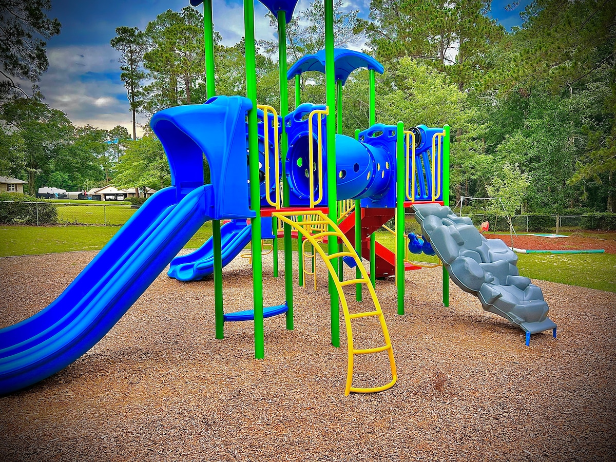 A colorful playground is showcased, featuring blue and yellow slides and climbing structures. The equipment is surrounded by a grassy area, providing ample space for children to play. Shafts of sunlight highlight the vibrant colors against a backdrop of trees.