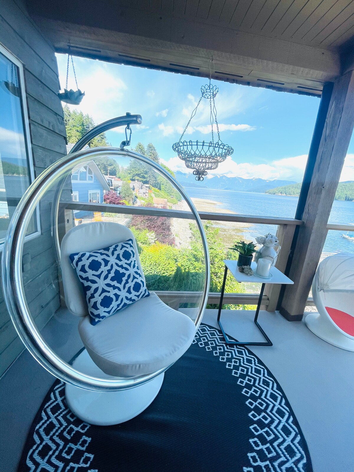 A modern hanging chair is positioned on the balcony, offering a view of the water and surrounding landscape. A small white table holds a potted plant, and a patterned rug lies beneath. Bright light fills the space, creating an inviting atmosphere.