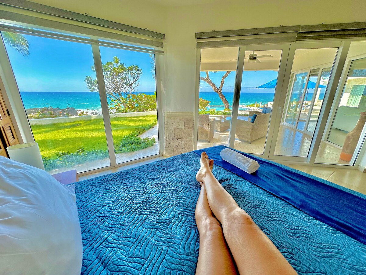 A spacious bedroom features a vibrant blue quilt on the bed, with large glass sliding doors revealing a stunning ocean view. Lush greenery is visible outdoors, and natural light brightens the room, creating a serene atmosphere.