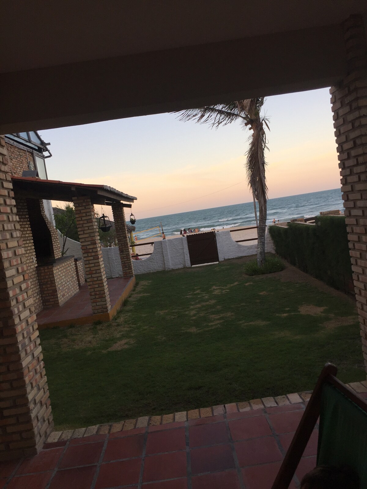 A view from a shaded porch reveals a peaceful beach scene. The sandy shore meets the calm ocean under a soft pastel sky. Lush green grass leads to a gated entrance, with palm trees swaying gently in the breeze, adding a touch of tropical ambiance.