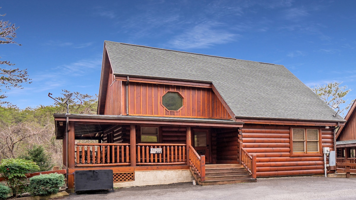 The cabin exterior is crafted from natural wood, showcasing a spacious porch with steps leading up to the entrance. A circular window is positioned prominently in the gabled wall, adding character to the rustic design.