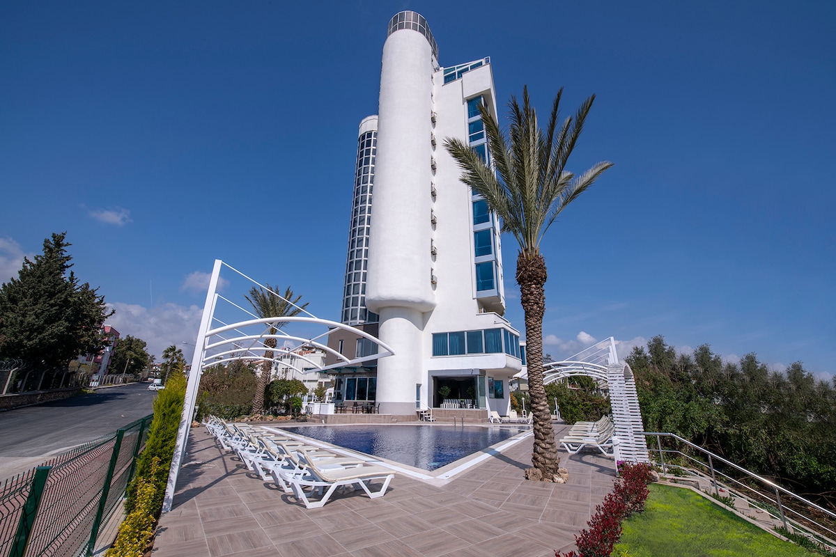 A modern multi-story building features a large glass façade and a cylindrical tower. An outdoor swimming pool is framed by lounge chairs and palm trees, while landscaped greenery surrounds the area, contributing to a relaxing environment.