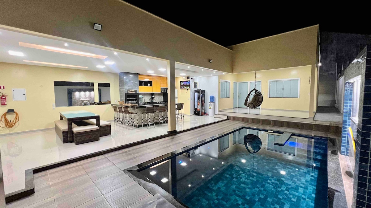 A spacious outdoor area features a modern pool with a clear surface reflecting surrounding lights. An adjacent bar area is equipped with high stools and appliances. A lounge chair hangs from the ceiling, adding a relaxing touch. Large windows allow ample natural light into the interior.