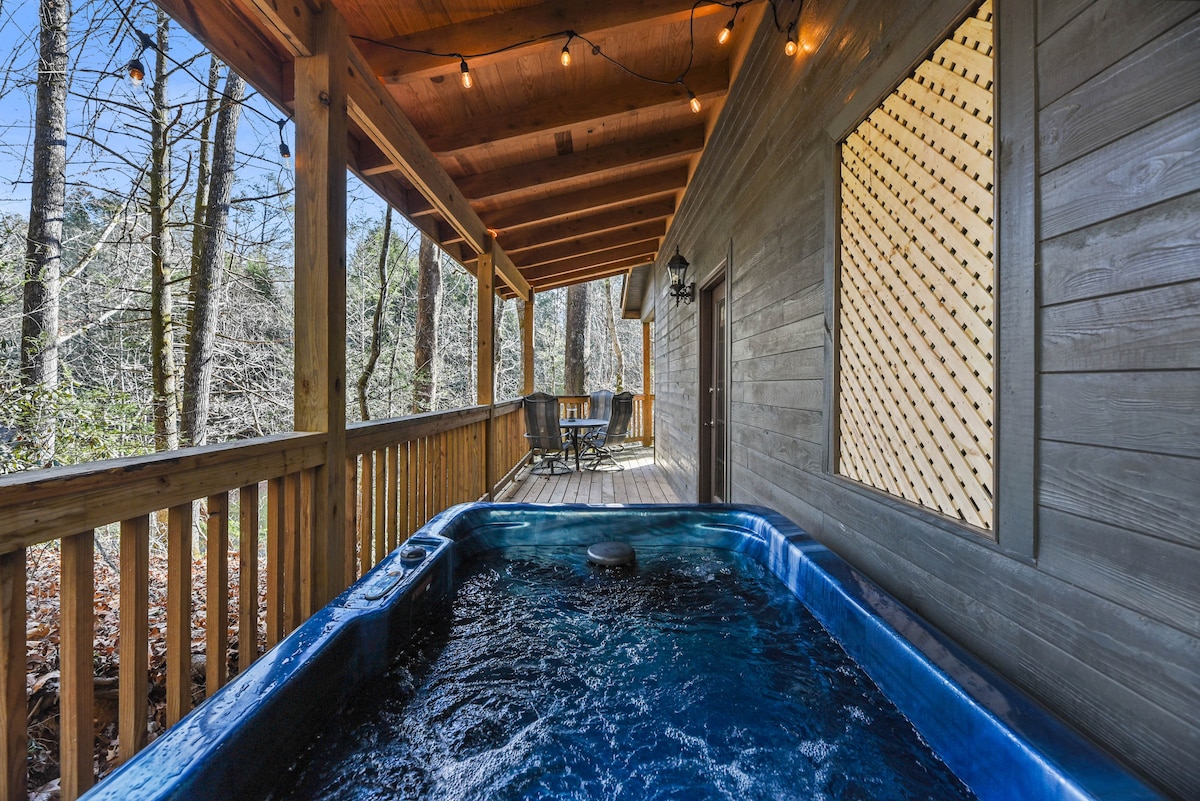 A private porch area features a hot tub, surrounded by wooden decking and a railing. String lights are suspended overhead, adding gentle illumination. Nearby, a small table and chairs are visible, offering a space for outdoor relaxation.
