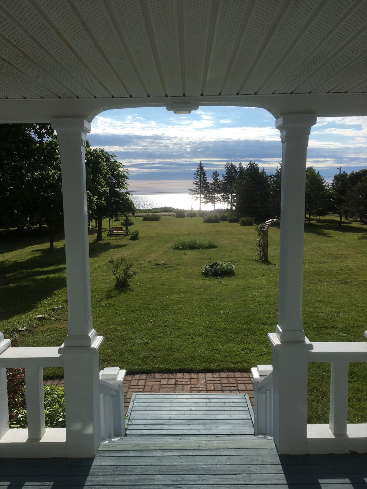 A view from a porch features greenery leading to the water, with grass and trees outlining the scenic landscape. The sunlight reflects off the water's surface, enhancing the calm ambiance. A path made of brick and wood is visible, leading to the open lawn.