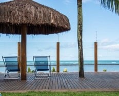 A shaded seating area features two reclining chairs beneath a thatched roof. The wooden deck extends over the water, offering views of the ocean. Palm trees provide a natural backdrop, enhancing the coastal atmosphere.