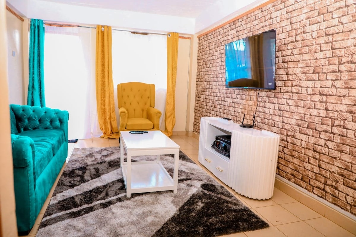A cozy living area is featured with a teal sofa and a bright yellow armchair. A white coffee table sits on a soft area rug, while a mounted television is visible on a white cabinet against a textured brick wall. Natural light streams in through large windows.