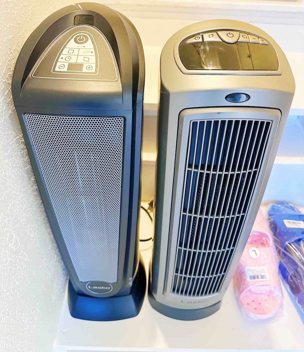 Two modern electric space heaters are positioned side by side, showcasing sleek designs in neutral tones. Each heater features control panels with buttons and a digital display. They are placed on a shelf with additional items visible in the background.