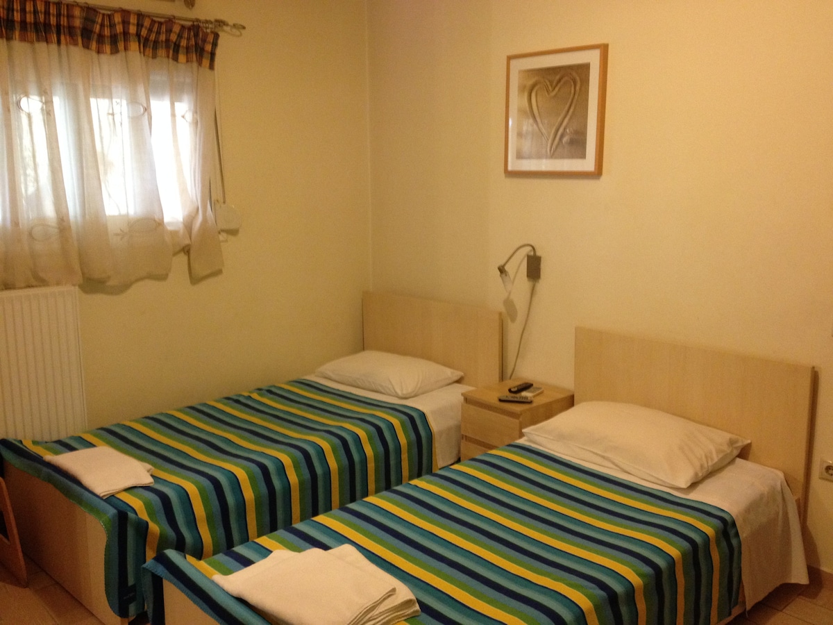The room contains two twin beds with striped blue and green bedding, positioned parallel to each other. A bedside table with a phone is located next to one bed. Natural light enters through a window, adorned with light drapes, illuminating the soft, neutral walls.