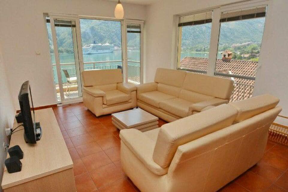 A light-filled living area is furnished with two beige sofas and a coffee table. Large windows provide views of the surrounding mountains and water. The space is enhanced by tiled flooring and a neutral colour palette, creating a comfortable and inviting environment.