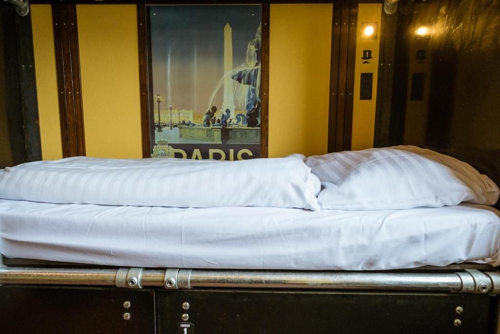 A neatly made single bed is presented with crisp white linens and pillows. A vintage-style poster of Paris hangs on the wall above, adding a touch of charm to the compact sleeping area.