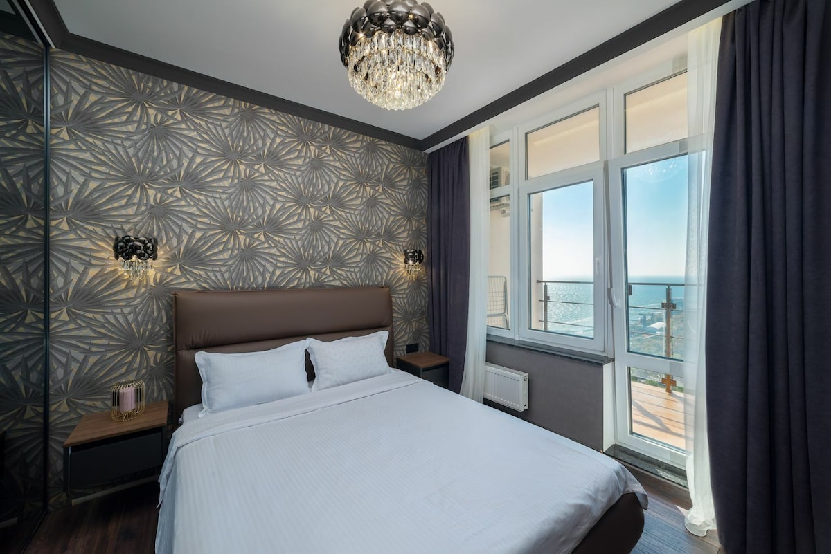 A well-appointed bedroom features a spacious bed with crisp white linens. Large windows provide a view of the sea, while elegant wallpaper and two light fixtures enhance the room's design. Curtains frame the windows, allowing for natural light to enter.