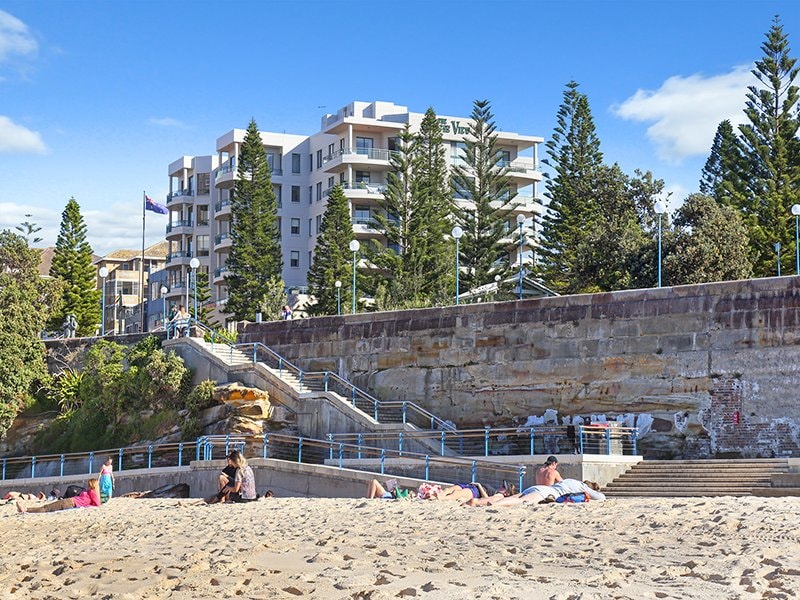 A view of Coogee Beach showcases soft sand with individuals lounging in the sun. In the background, the apartment building features modern architecture framed by tall pine trees. Clear skies enhance the overall ambiance of the beach, while a staircase leads up to the promenade.