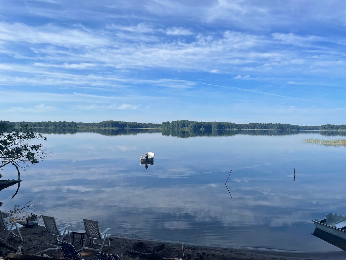 A serene lake scene is captured, featuring calm waters reflecting the sky and surrounding trees. A small boat is anchored nearby, while several deck chairs are positioned on the sandy shore, providing a tranquil setting for relaxation.