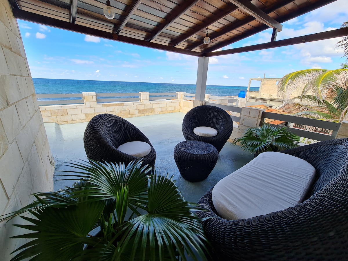 A shaded outdoor seating area features dark wicker chairs surrounding a small table. Lush green plants are positioned around the space, with the ocean visible in the background. The sky above is partly cloudy, enhancing the serene coastal atmosphere.