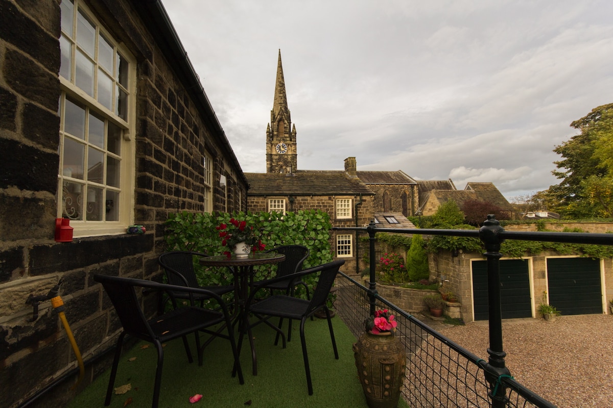 A balcony with a small table and four chairs is shown, surrounded by flowers in pots. The view includes a historic church spire and stone buildings, creating a charming atmosphere. The balcony features artificial grass, enhancing the outdoor space.