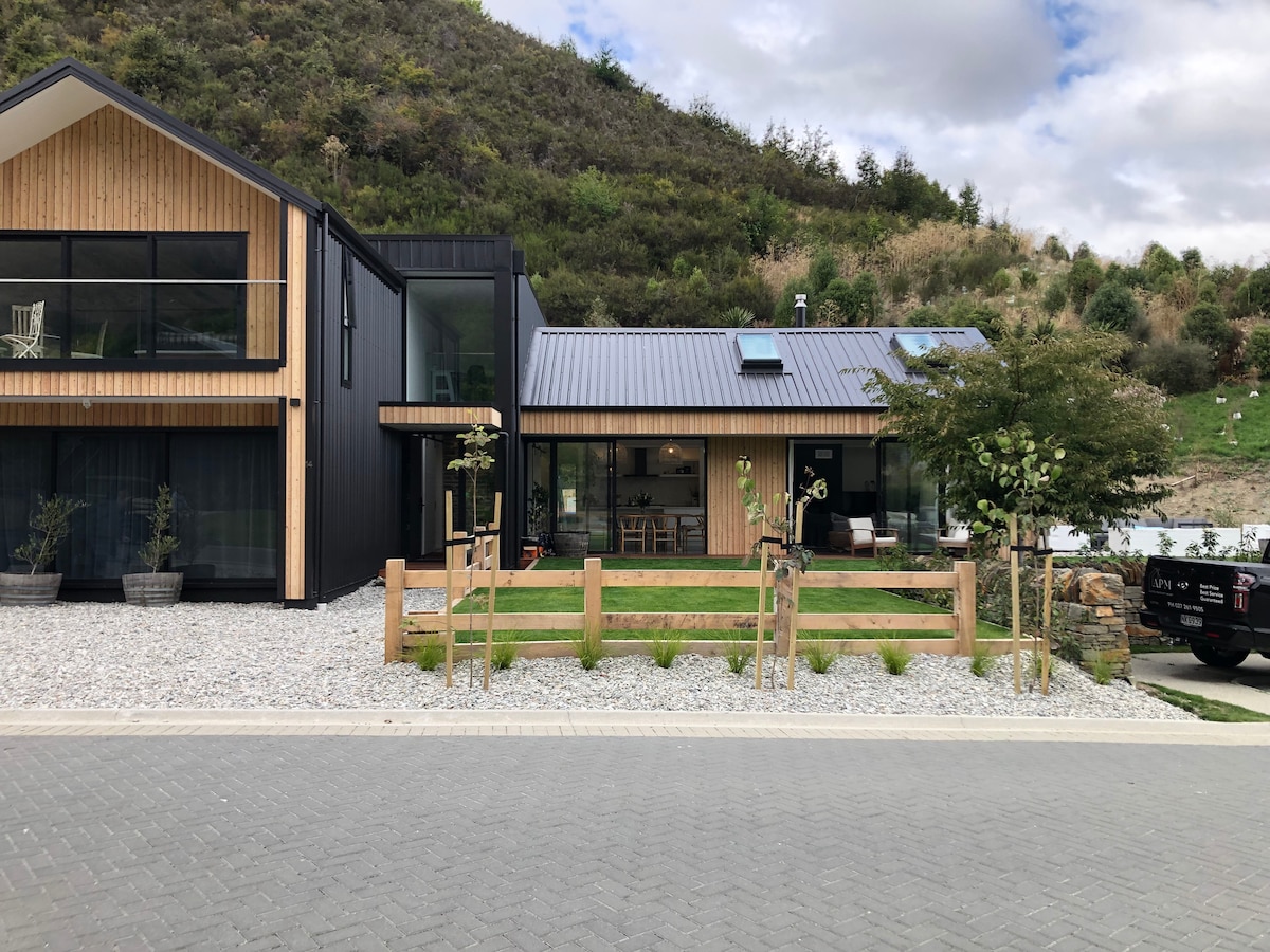 The exterior of a modern home is displayed, featuring a combination of wood and dark cladding. A grassy area with small trees and a wooden fence is visible in the foreground. Stone pathways and gravel create a welcoming entryway, set against a backdrop of gentle hills.