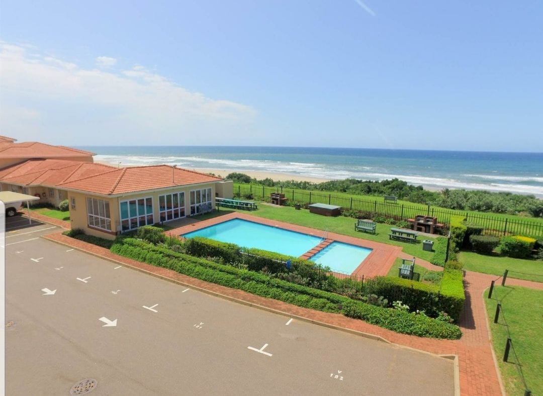 An outdoor pool is situated beside the ocean, surrounded by well-maintained greenery and lounge chairs. The pool area is framed by a paved pathway and offers views of the ocean and distant shoreline. A colorful sky adds to the serene setting.