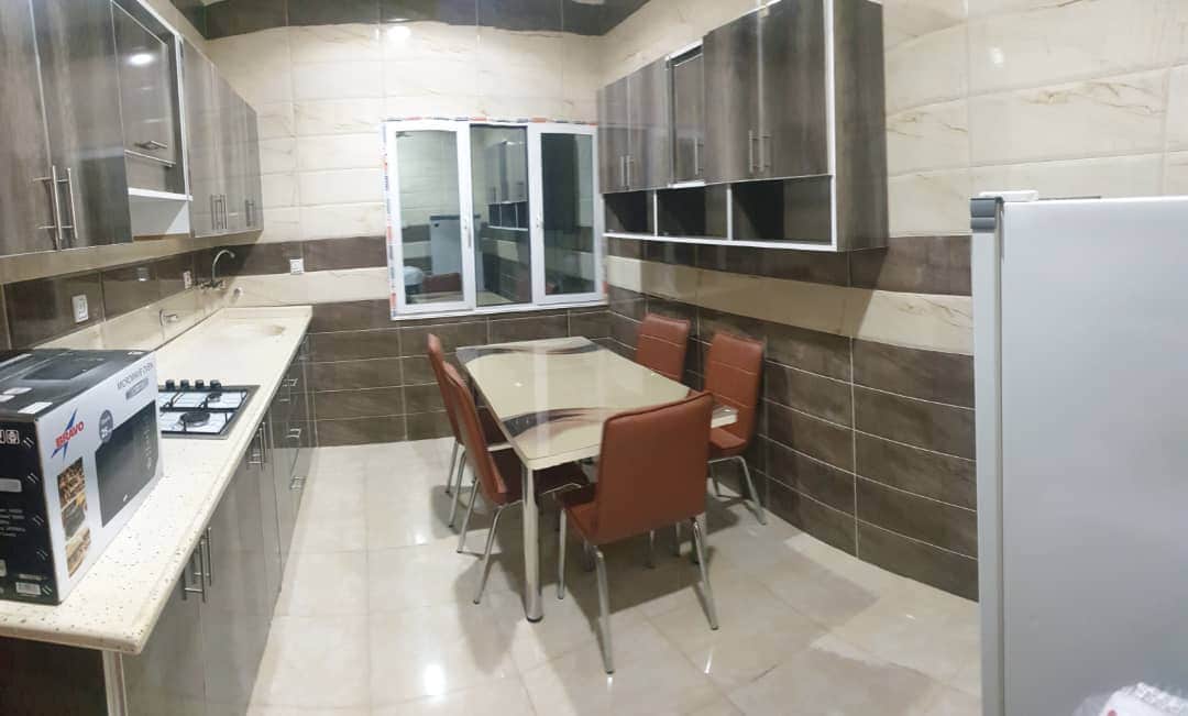 A fully equipped kitchen features modern cabinetry and a smooth countertop. A table with five chairs sits at the center, providing an inviting space for dining. A large window offers natural light, and essential appliances are visible, including a gas stove and a refrigerator.