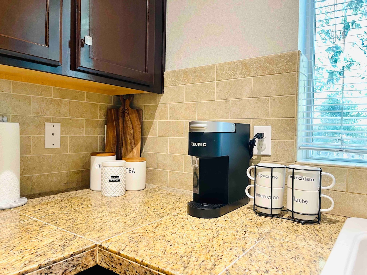 A kitchen corner is depicted with a Keurig coffee maker situated on a textured granite countertop. Container jars labeled for sugar, tea, and coffee are arranged nearby, along with four ceramic mugs displayed on a metal rack. Natural light filters through a window, illuminating the workspace.