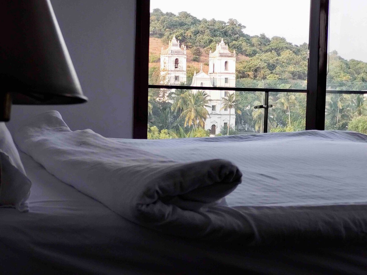 A well-structured bed is neatly arranged with white linens, showcasing a rolling pillow. Through the large window, views of lush greenery and a historic church on a hill can be observed, allowing natural light to fill the room.
