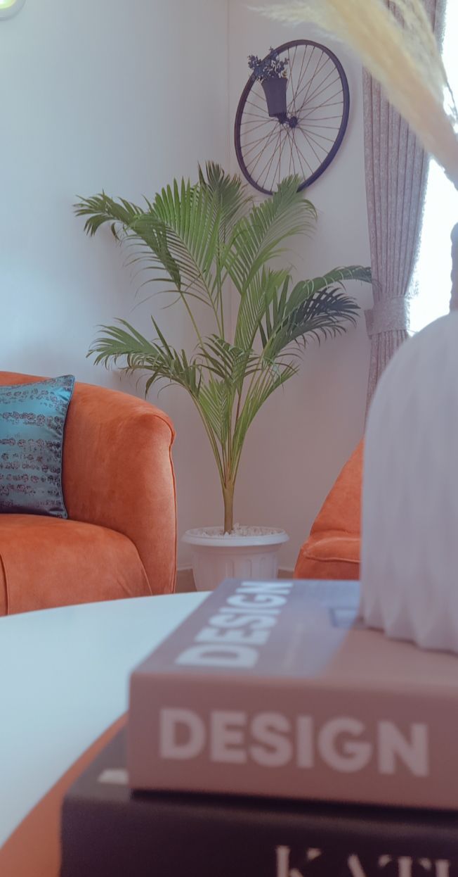 A cozy seating area is highlighted by two orange chairs with decorative cushions. A decorative plant is positioned on a white pedestal in the background, while a stylish coffee table is visible in the foreground, adorned with books that feature modern design themes.