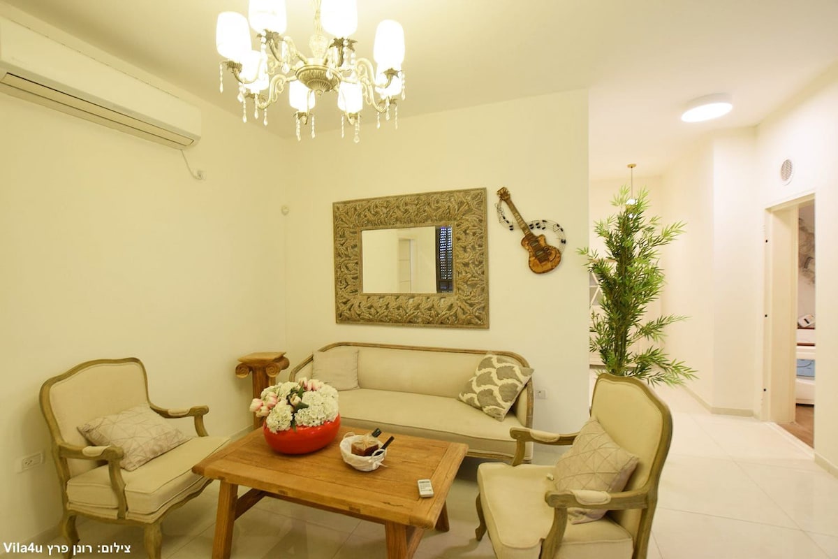 A cozy seating area is furnished with two elegant armchairs and a wooden table, topped with a decorative bowl of flowers. A large mirror reflects the room's light, complemented by a soft ceiling chandelier. A tall plant adds a touch of greenery to the space.