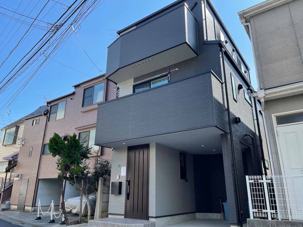 A modern three-story house is depicted from the street view, featuring a sleek exterior with a combination of light and dark grey panels. Large windows are visible on each level, along with a minimalist entrance framed by an elegant door. Surrounding greenery complements the contemporary design.