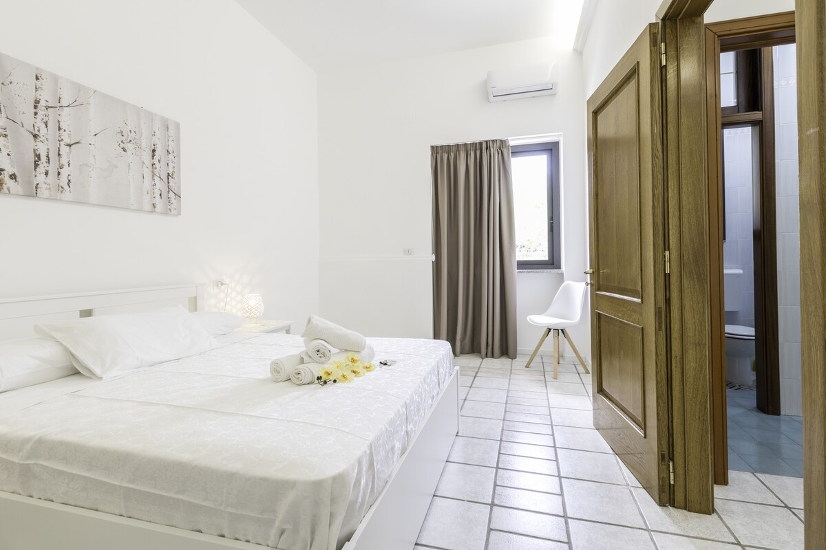 A bright bedroom features a spacious bed with two neatly arranged towels and decorative flowers. A window allows natural light to brighten the room, and a door leads to a private bathroom. Neutral walls and tiled flooring contribute to a clean and open atmosphere.