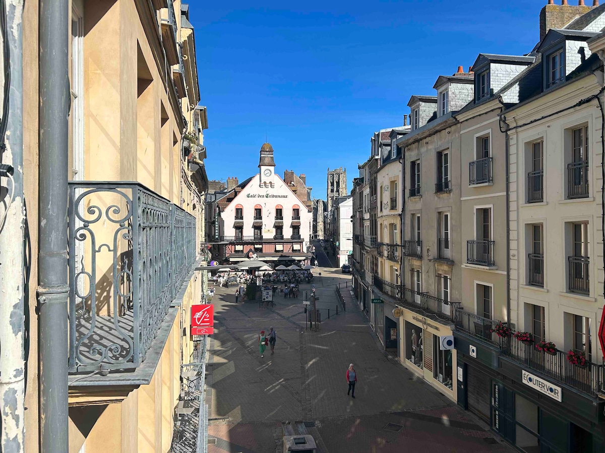 A view from a second-floor balcony showcases an open square bustling with activity. The iconic Café des Tribunaux is centrally positioned, with its distinct architecture and outdoor seating inviting visitors. Stone buildings line the square, their facades displaying a blend of colors under clear blue skies.