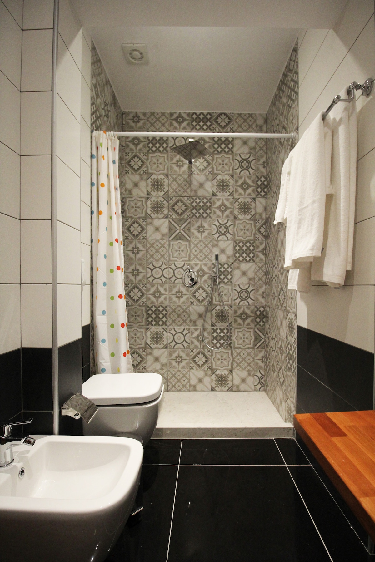 A modern bathroom features intricate patterned tiles on the walls, complementing a sleek shower area. Towels are neatly hung on a rack, and the space includes a bidet and a white toilet. Natural light illuminates the room, highlighting the contemporary design.