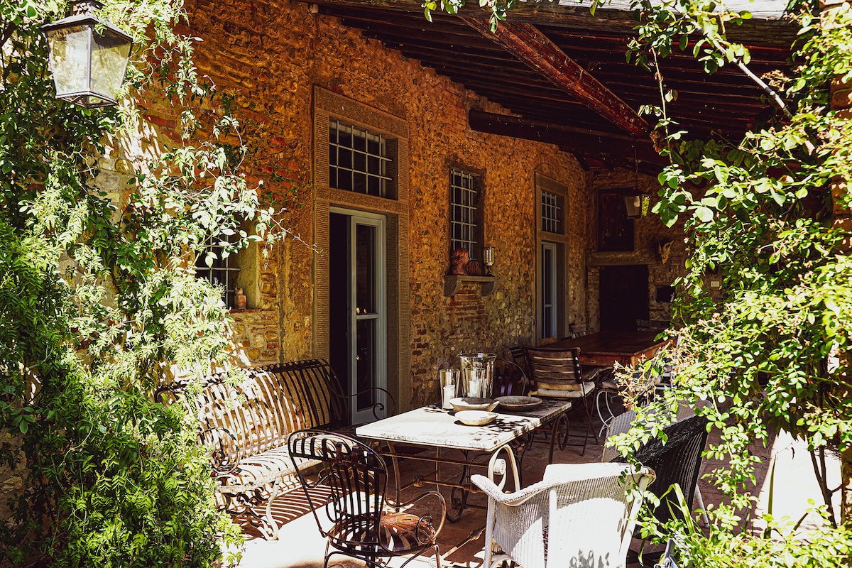 An outdoor patio area is framed by lush greenery, featuring a combination of wrought iron and wooden furniture. A table is set for dining, complemented by various seating options. The rustic stone walls of the farmhouse provide a charming backdrop.