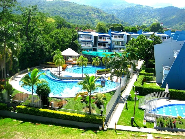 The outdoor area features multiple swimming pools surrounded by landscaped gardens. Lush palm trees provide shade, and lounge areas are available for relaxation. The backdrop consists of multi-story buildings nestled against a scenic mountain landscape under a clear sky.