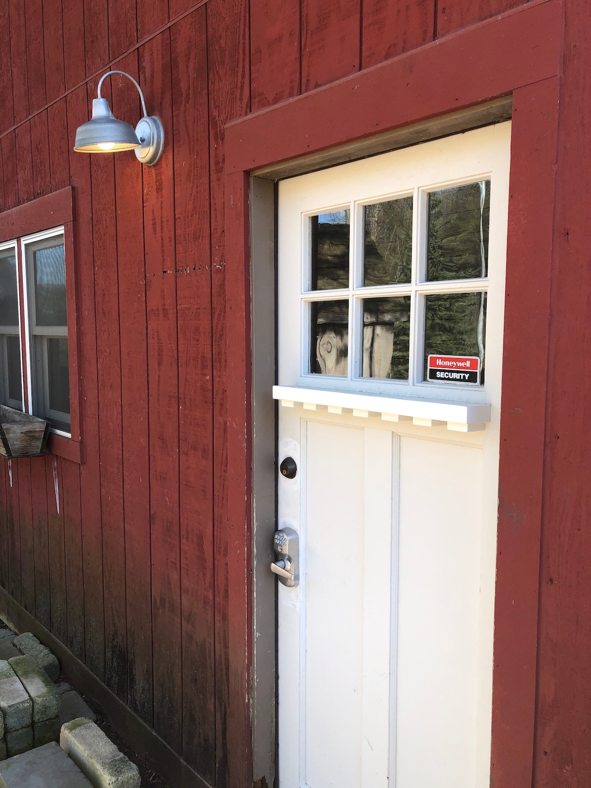 The entrance features a sturdy white door with four square windows on the upper section, allowing some light to enter. The door is complemented by a small overhang and a wall-mounted light fixture. The red wooden siding shows weathered textures, adding character to the structure.