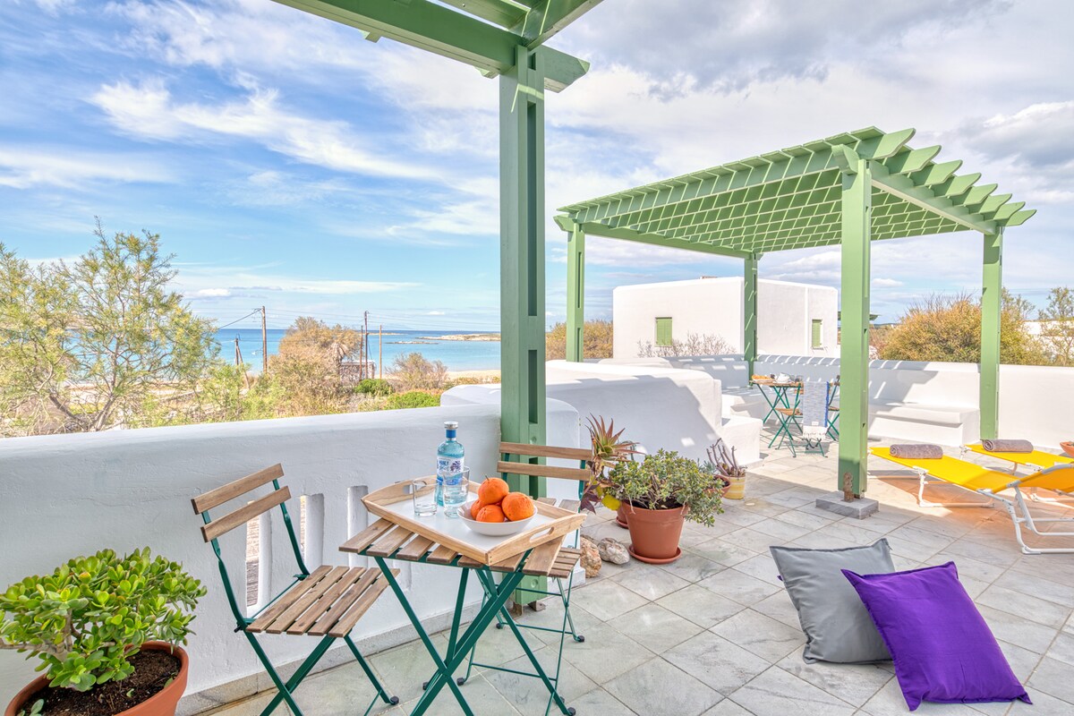 A spacious terrace features a green pergola providing shade. A small dining table set for two is visible, accompanied by potted plants and a colorful pillow. The serene views of the ocean and surrounding greenery can be seen in the background, enhancing the outdoor atmosphere.