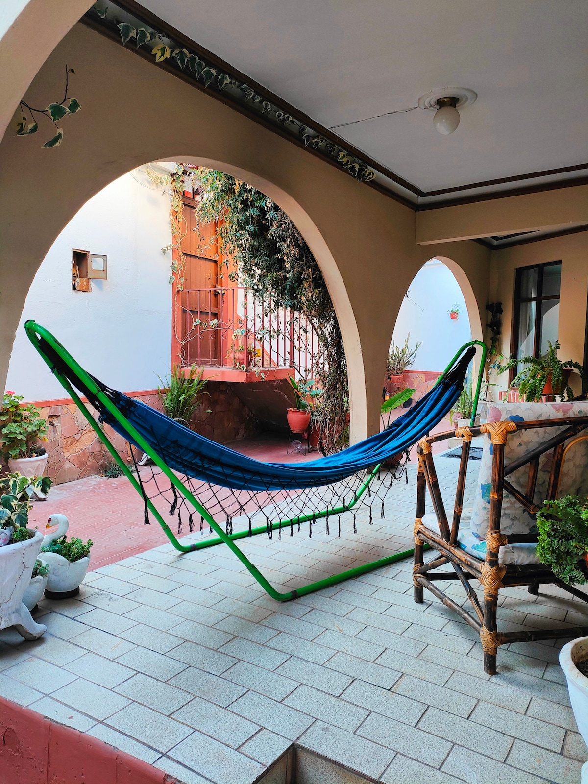 A hammock in vibrant blue hangs in a shaded outdoor area, surrounded by potted plants. Arched walls frame the space, while tiled flooring adds a touch of elegance. Natural light filters gently through the open entrance, creating a calm and inviting atmosphere.