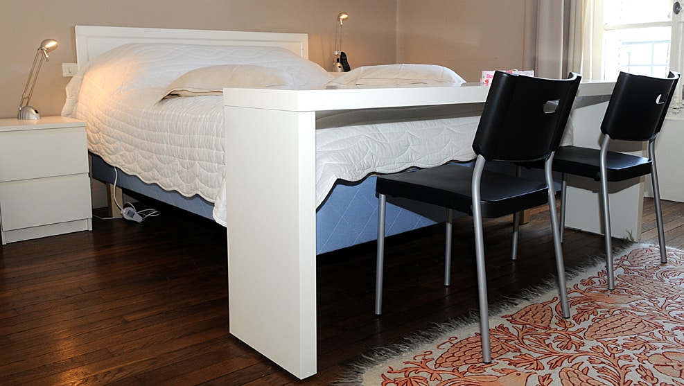 A spacious bed is positioned against a neutral wall, accompanied by two modern chairs at a minimalist table. A warm wooden floor enhances the room's inviting atmosphere, while natural light filters through a nearby window, illuminating the space.