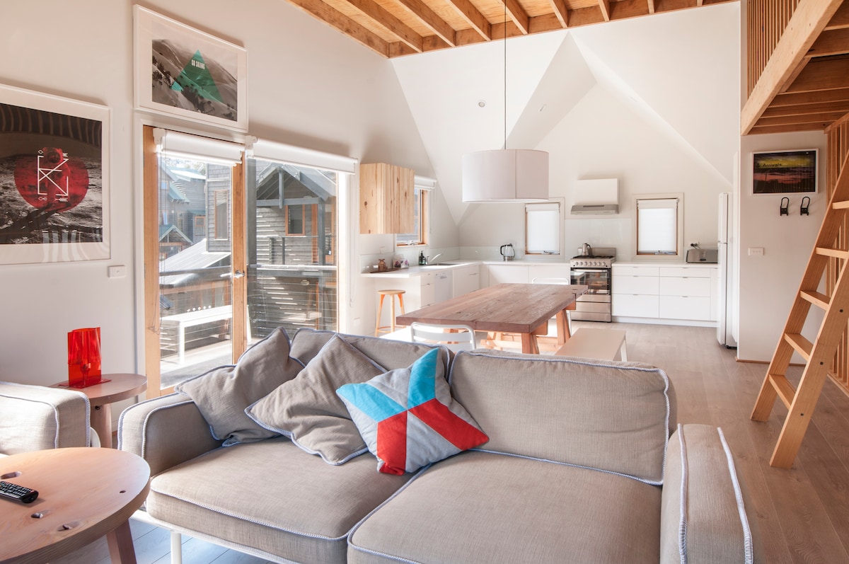 An open living space features a cozy sofa adorned with colorful cushions, facing a large wooden dining table. The modern kitchen is equipped with sleek cabinets and appliances. Large windows allow natural light to illuminate the room, enhancing the airy atmosphere.