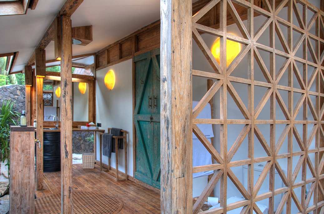 A natural wooden structure features an open layout with a patterned wooden screen, creating a sense of privacy. Soft pendant lights provide ambient illumination. Modern amenities are visible, including a sink and towel rack, seamlessly integrated into the rustic design.