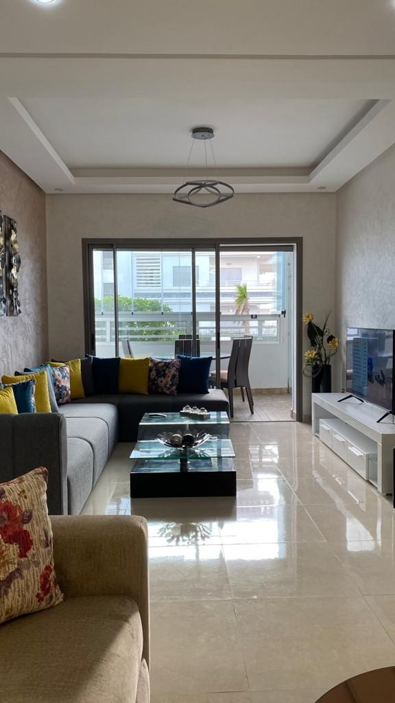 A bright living area features a sectional sofa adorned with colorful cushions, complemented by a sleek coffee table at its center. Large windows provide natural light, with a balcony view outlined by indoor plants. A television is positioned on a modern cabinet.