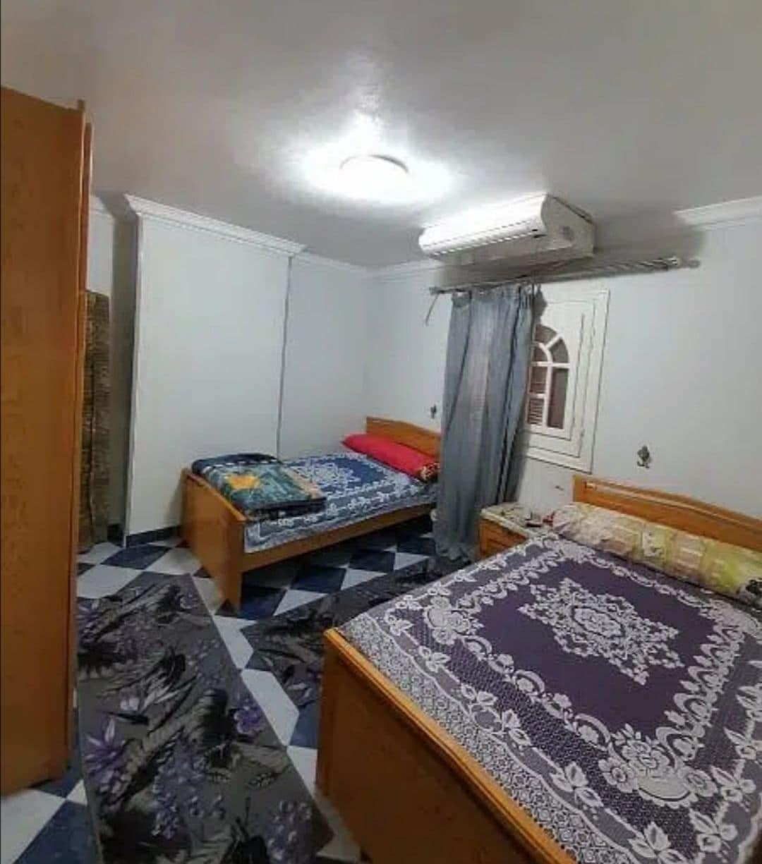 The sleeping area features two spacious beds, each with distinct patterned bedding in shades of purple and blue. A light fixture illuminates the room, while an air conditioning unit is mounted on the wall. A patterned rug complements the tiled floor.
