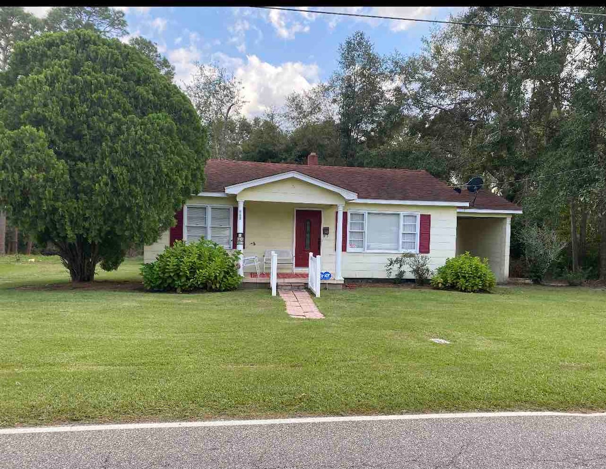 The exterior view showcases a single-story home with yellow siding and red shutters. A well-maintained lawn is present, framed by lush green shrubs. A welcoming front porch is visible, and a large tree stands beside the house, offering shade to the property.