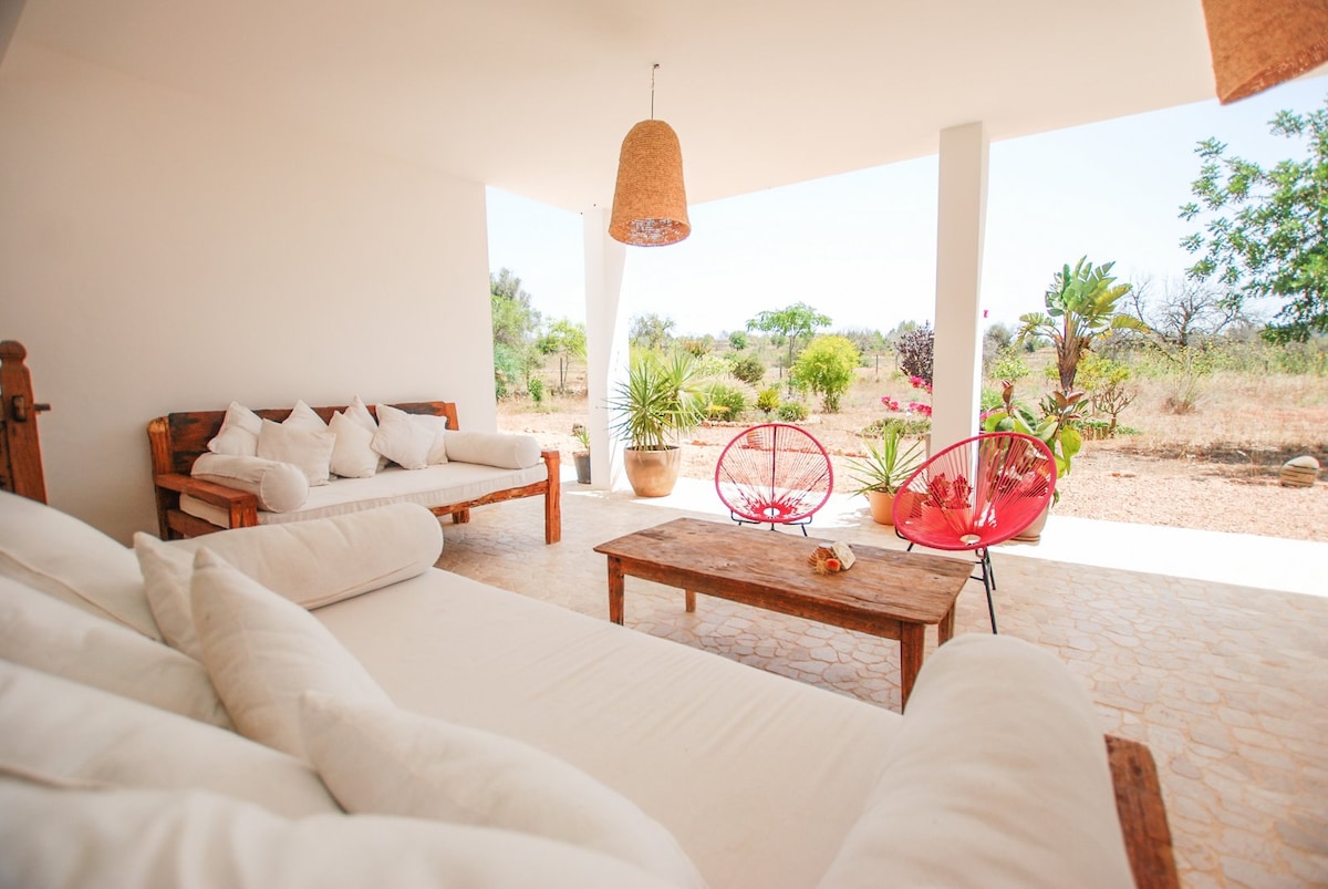 An outdoor seating area features a spacious wooden sofa adorned with light-colored cushions. Two vibrant red chairs complement the space under a pendant light, surrounded by lush greenery and natural landscape in the background, providing a serene and inviting setting.