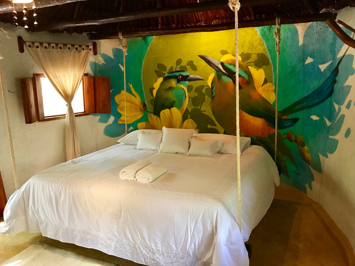 A spacious hanging bed is centrally located, complemented by soft white linens. A vibrant mural depicting two colorful birds adorns the wall behind the bed, surrounded by floral elements. Natural light filters through the window draped with sheer curtains, enhancing the room's inviting atmosphere.